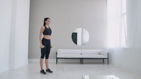 A-woman-raises-her-arms-with-dumbbells-doing-a-shoulder-exercise-in-her-light-white-apartment-against-a-sofa-and-window.